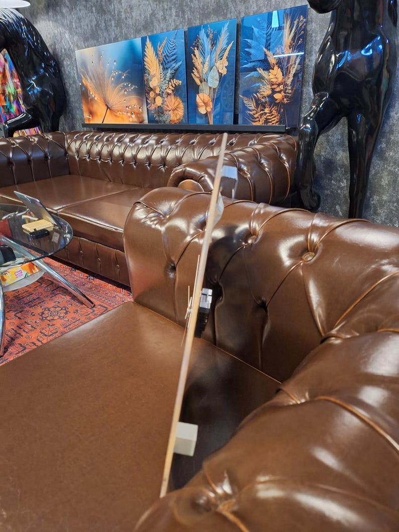 May include: A thin, flat, rectangular wall clock with a light-colored wooden face and black clock hands, positioned vertically on the arm of a brown tufted leather sofa. The clock mechanism is visible on the back side of the wooden panel.