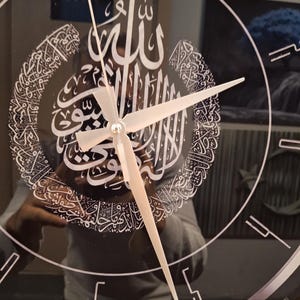 May include: A circular wall clock with a reflective black surface featuring white Arabic calligraphy in a crescent shape. The clock has thin white hour markers and simple, straight white hands. The design is centered on the clock face.