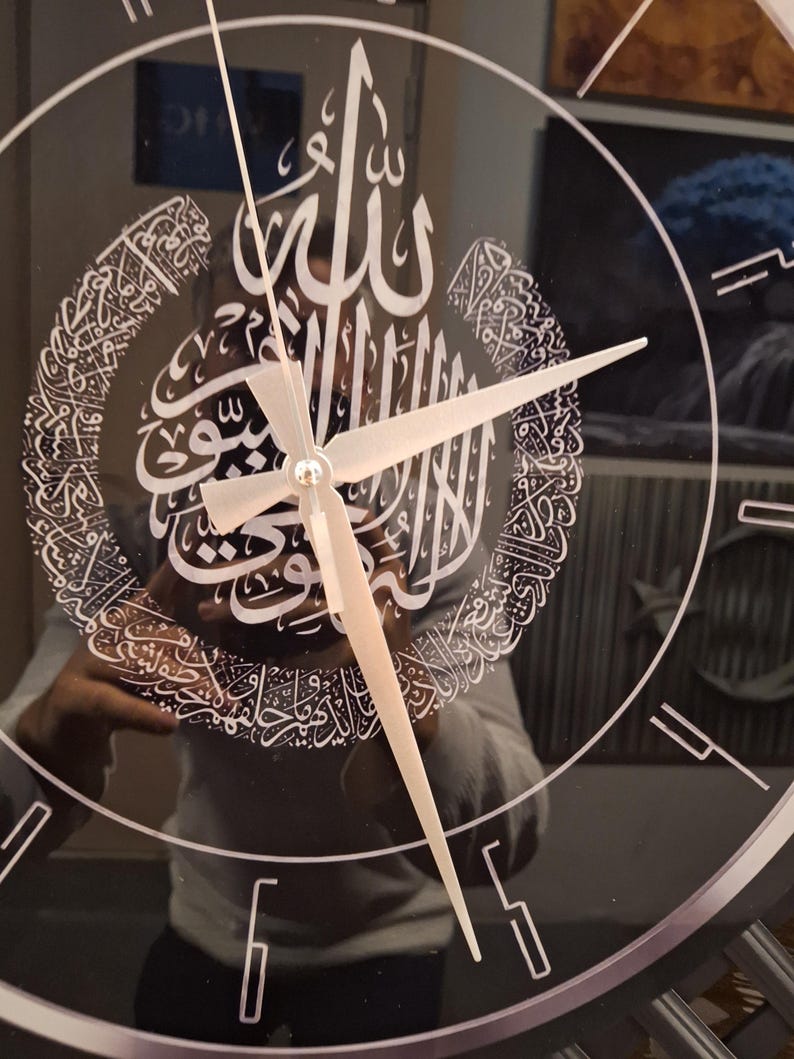 May include: A circular wall clock with a reflective black surface featuring white Arabic calligraphy in a crescent shape. The clock has thin white hour markers and simple, straight white hands. The design is centered on the clock face.