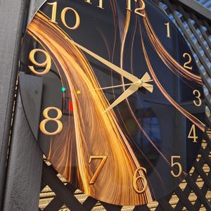 May include: A round wall clock with a black face featuring a swirling gold and amber abstract pattern. The clock has gold-colored numerals from 1 to 12 and matching gold-colored hour, minute, and second hands centered on the dial.