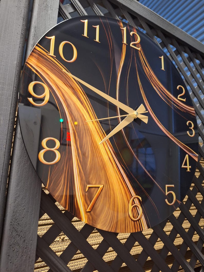 May include: A round wall clock with a black face featuring a swirling gold and amber abstract pattern. The clock has gold-colored numerals from 1 to 12 and matching gold-colored hour, minute, and second hands centered on the dial.
