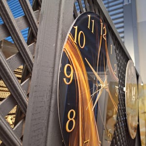 May include: A circular wall clock featuring a dark background with abstract golden light streaks. The clock face displays large, gold-colored Arabic numerals from 1 to 12 and thin, gold-colored hour, minute, and second hands.