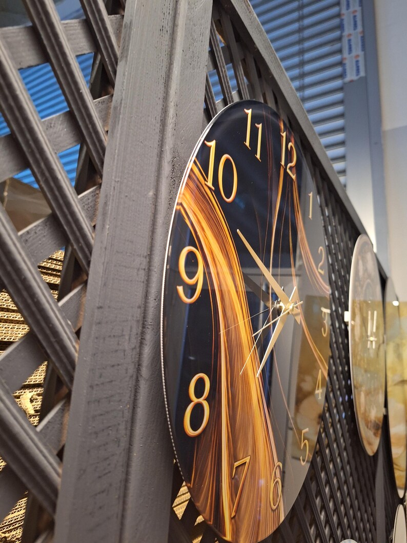 May include: A circular wall clock featuring a dark background with abstract golden light streaks. The clock face displays large, gold-colored Arabic numerals from 1 to 12 and thin, gold-colored hour, minute, and second hands.