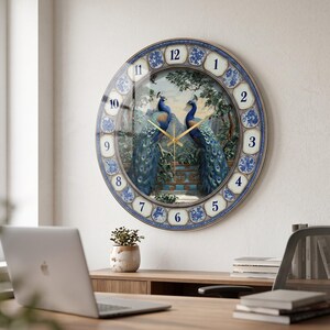 May include: A round wall clock featuring a central illustration of two peacocks facing each other amidst trees and a stone structure. The clock face has a blue and white floral patterned border with large, clear numbers from 1 to 12 and gold hands.