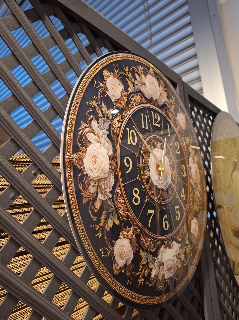 May include: A round wall clock featuring a dark blue background with gold-colored numerals and ornate gold filigree patterns. The clock face is decorated with several cream-colored rose illustrations and has thin gold-colored clock hands.