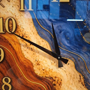 May include: A wall clock featuring a face with a swirling pattern of deep blue, tan, and dark brown wood grain textures. The clock has large gold-colored numerals from 7 to 12 and two thin black hands pointing toward the ten and twelve positions.
