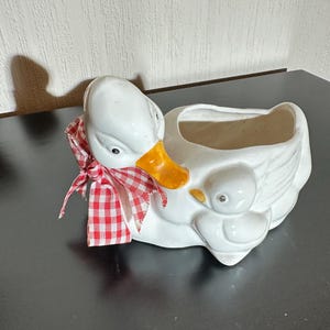 Puede incluir: Jardinera de cerámica blanca con forma de pato, pico naranja y lazo a cuadros rojos y blancos. La jardinera presenta un patito en un lateral. Diseñada para plantas o flores.