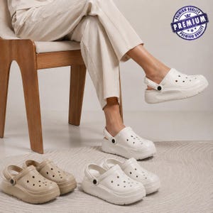 May include: White and beige platform clogs featuring a perforated upper, a rounded toe, and a pivoting heel strap with a black circular rivet. The footwear has a thick, moulded sole and a smooth finish. A blue circular badge with the text PREMIUM is visible.