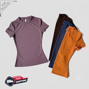 May include: A collection of four short-sleeved crew neck t-shirts in solid colors. The shirts are displayed in a stack, featuring a mauve shirt in the foreground, followed by brown, navy blue, and orange shirts layered behind it on a plain background.