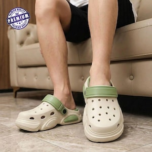 May include: A pair of off-white clogs with a sage green heel strap and side accents. The footwear features a perforated toe box, a thick sole with moulded detailing, and a circular blue and white badge in the corner reading Premium Quality.