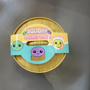 May include: A yellow, round toy with a woven texture, featuring a label that reads "Squishy Dumpling." The label has cartoon dumpling characters and the word "Squeeze It!" The toy is designed to be squeezed.