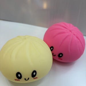 May include: Two round, cartoon-style toys resembling dumplings. One is pale yellow, the other is bright pink. Both have smiling faces with black eyes and rosy cheeks. The toys are on a white surface.