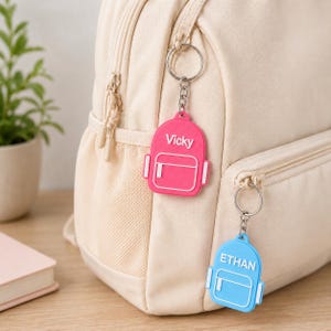 May include: Two backpack-shaped keychains attached to a beige backpack. One keychain is hot pink with the white text Vicky, and the other is light blue with the white text ETHAN. Both feature a metal ring and chain, with white outlines of a pocket.