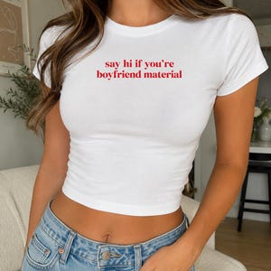 May include: A white short-sleeved crew neck t-shirt featuring the red text "say hi if you're boyfriend material" printed across the chest. The garment is a fitted, cropped style paired with light blue denim jeans with a visible metal button.