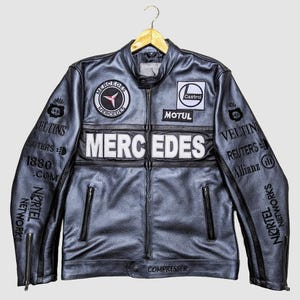 May include: A black leather jacket with multiple patches and logos, including "MERCEDES" in large white letters across the chest. Additional logos include "Castrol" and "MOTUL". The sleeves feature text and logos, and the jacket has a zipper closure.