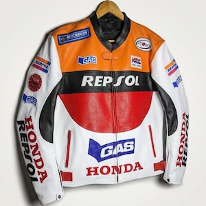 Puede incluir: Chaqueta de moto de cuero blanco, naranja, negro y rojo, con logotipos de patrocinadores como "Repsol", "Honda" y "Michelin". La chaqueta está colgada de una percha de madera. Tiene cuello y puños negros.