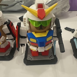Pu&ograve; includere: Una collezione di figure in stile Gundam stampate in 3D e colorate. La figura centrale &egrave; un robot bianco, rosso, giallo, blu e verde con una pistola. Le figure sono su una superficie bianca, con altre parti di robot nelle vicinanze.