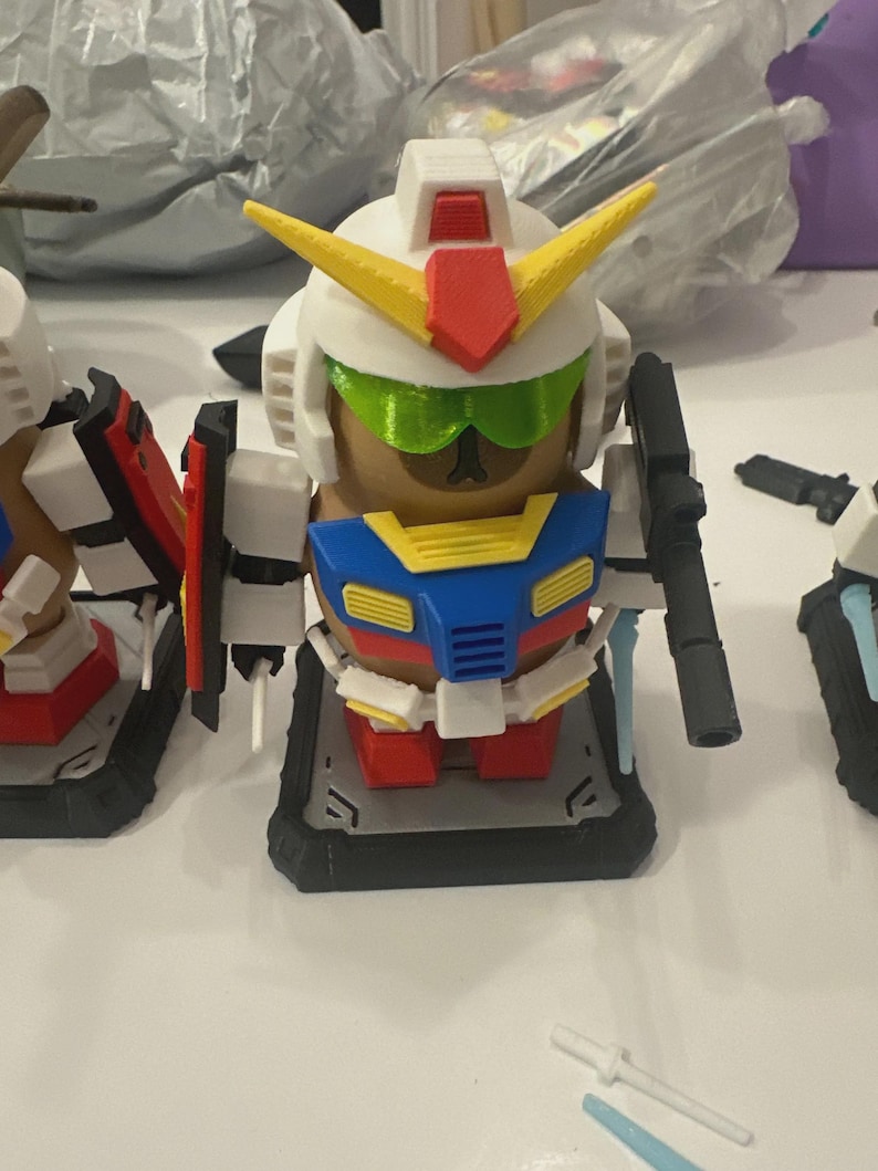Pu&ograve; includere: Una collezione di figure in stile Gundam stampate in 3D e colorate. La figura centrale &egrave; un robot bianco, rosso, giallo, blu e verde con una pistola. Le figure sono su una superficie bianca, con altre parti di robot nelle vicinanze.