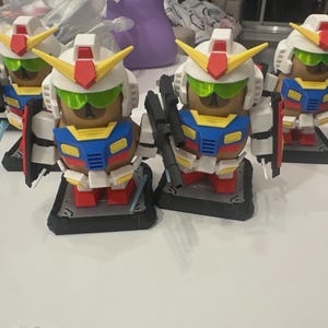 Pu&ograve; includere: Quattro figurine in stile Gundam con facce marroni, elmetti bianchi e armature colorate. Ogni figura ha uno scudo, un'arma e poggia su una base nera. Gli elmetti presentano dettagli gialli e rossi e visiere verdi.