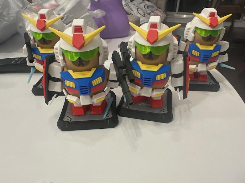 Pu&ograve; includere: Quattro figurine in stile Gundam con facce marroni, elmetti bianchi e armature colorate. Ogni figura ha uno scudo, un'arma e poggia su una base nera. Gli elmetti presentano dettagli gialli e rossi e visiere verdi.