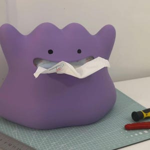 Puede incluir: Una caja de pañuelos morada con forma de personaje de dibujos animados, con una cara sonriente y pañuelos saliendo de su boca. La caja está sobre una esterilla de corte verde, con herramientas cerca.