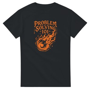 Könnte beinhalten: Ein schwarzes T-Shirt mit kurzen Ärmeln und einer orangefarbenen Grafik auf der Brust. Das Design zeigt den Text "PROBLEM SOLVING 101" über einer Illustration eines brennenden Kometen oder Feuerballs, umgeben von kleinen vierzackigen Sternen.