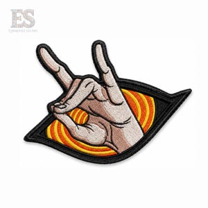 Op de afbeelding: Een geborduurde patch met een hand die een rock-and-roll-gebaar maakt, geplaatst over een geel en oranje spiraalontwerp. De hele afbeelding is ingesloten in een zwarte, ruitvormige rand met dikke, donker gestikte randen.