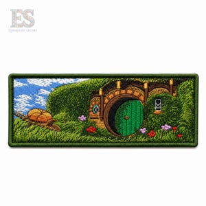 May include: A rectangular embroidered patch featuring a landscape with a round green door set into a grassy hill. The scene includes a wooden wheelbarrow with a pumpkin, a blue sky with white clouds, and small red and purple flowers in the grass.