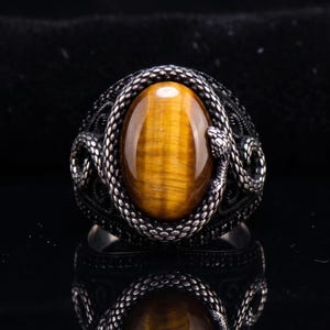 Peut inclure: Bague argentée ornée d'une pierre œil de tigre ovale. La bague présente un motif de serpent détaillé autour de la pierre et sur le bandeau. La pierre œil de tigre est de couleur brun doré chaud.