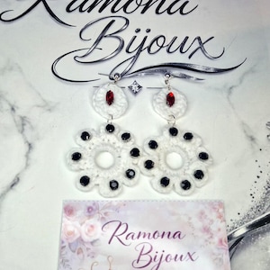 Könnte beinhalten: Weiße Häkelohrringe mit schwarzen und roten Akzenten. Die Ohrringe sind blütenförmig mit einem weißen Mittelkreis und schwarzen Perlen an den Rändern. Das Bild zeigt auch den Text "Ramona Bijoux" und "HANDMADE CROCHET".