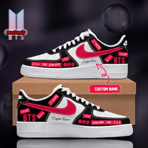 Op de afbeelding: Een paar wit-zwarte sneakers met rode Nike swooshes en rode accenten. De schoenen zijn versierd met de tekst BTS, J-HOPE, SUGA, RM, JIN, JIMIN en JUNG KOOK. Een tijdelijke aanduiding voor een aangepaste naam is zichtbaar op de zijkant.