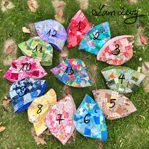Op de afbeelding: Een verzameling van dertien patchwork emmerhoeden in verschillende kleuren en patronen, waaronder rood, blauw, groen en geel. Elke hoed is genummerd en wordt getoond op groen gras. De hoeden zijn gemaakt van stof.