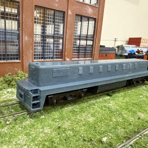 Può includere: Una locomotiva di treno in miniatura di colore grigio con un corpo lungo e rettangolare dotato di prese d'aria e pannelli laterali. Il motore poggia su ruote nere e binari metallici davanti a un edificio in mattoni con grandi finestre a griglia.