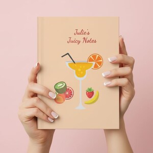 May include: A notebook with the text "Julie's Juicy Notes" and a cocktail illustration. The cover features a margarita glass, orange slice, kiwi, strawberry, and banana. The notebook is held in front of a pink background.