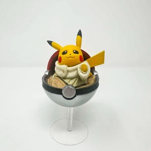 以下が含まれることがあります： 黒い耳と赤い頬を持つ黄色のピカチュウのフィギュア。銀と黒のモンスターボールの中。透明なスタンドに置かれた、ユニークなコレクターズアイテムです。