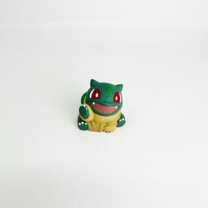 May include: A green and yellow Bulbasaur figurine with red eyes and a smiling expression. The figure is a stylized representation of the popular Pok&eacute;mon character, with a detailed design and a smooth finish. The figurine is set against a plain white background.