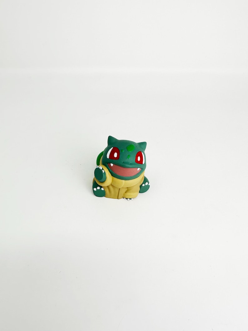 May include: A green and yellow Bulbasaur figurine with red eyes and a smiling expression. The figure is a stylized representation of the popular Pok&eacute;mon character, with a detailed design and a smooth finish. The figurine is set against a plain white background.