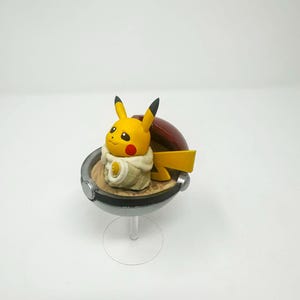 以下が含まれることがあります： 黒い先端の耳と赤い頬を持つ黄色のピカチュウのフィギュアが、赤と銀色のモンスターボールの中に座っています。 このフィギュアは透明なスタンドにあり、人気のポケモンフランチャイズの収集品を紹介しています。