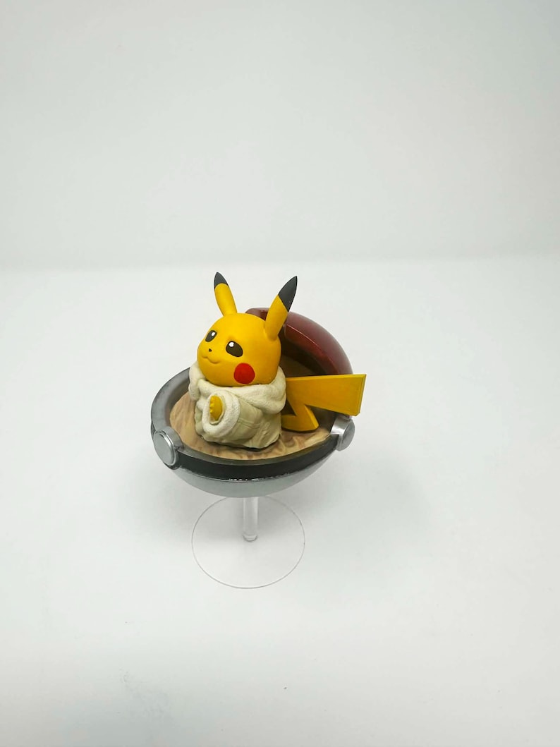 以下が含まれることがあります： 黒い先端の耳と赤い頬を持つ黄色のピカチュウのフィギュアが、赤と銀色のモンスターボールの中に座っています。 このフィギュアは透明なスタンドにあり、人気のポケモンフランチャイズの収集品を紹介しています。