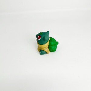 May include: A green Bulbasaur figurine with red eyes and a yellow collar. The figurine has a large green bulb on its back. The Bulbasaur is a popular character from the Pokemon franchise.