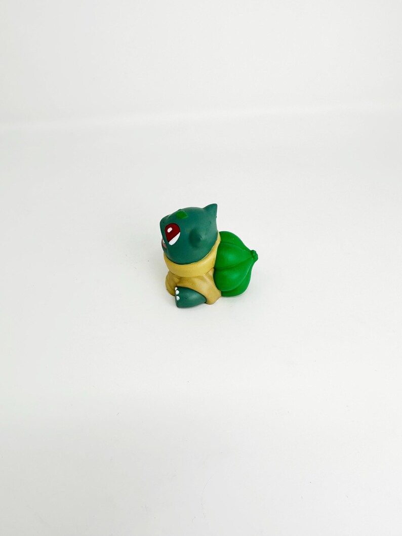 May include: A green Bulbasaur figurine with red eyes and a yellow collar. The figurine has a large green bulb on its back. The Bulbasaur is a popular character from the Pokemon franchise.