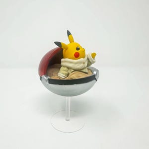 以下が含まれることがあります： クリーム色の衣装を着た黄色いピカチュウのフィギュアが、赤と銀色のモンスターボールの中に座っています。フィギュアは透明なスタンドに取り付けられており、ポケモンフランチャイズのコレクターアイテムです。