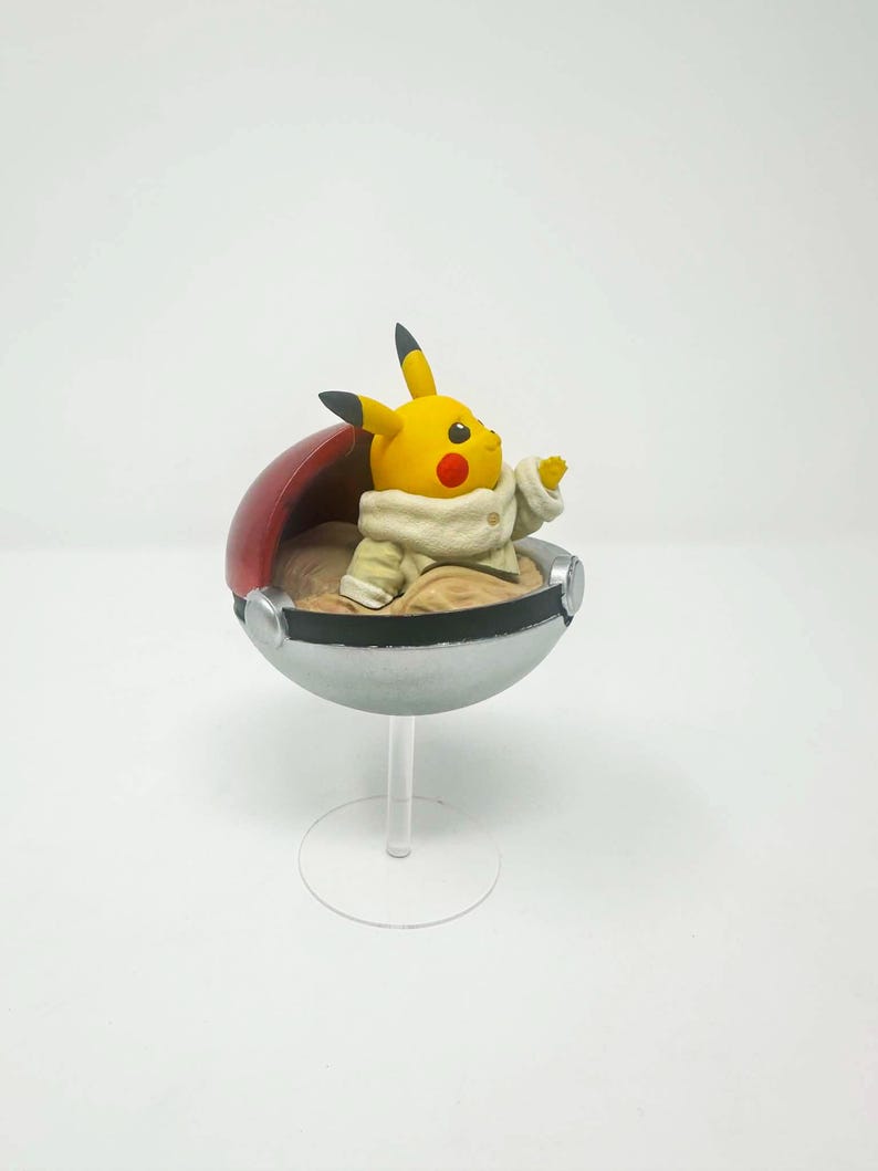 以下が含まれることがあります： クリーム色の衣装を着た黄色いピカチュウのフィギュアが、赤と銀色のモンスターボールの中に座っています。フィギュアは透明なスタンドに取り付けられており、ポケモンフランチャイズのコレクターアイテムです。