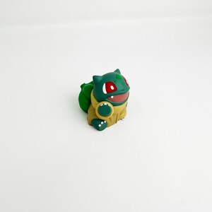 May include: A green and yellow Bulbasaur figurine with red eyes and a smiling mouth. The figurine has a large green bulb on its back and is set against a white background. The Bulbasaur is a popular character from the Pokemon franchise.