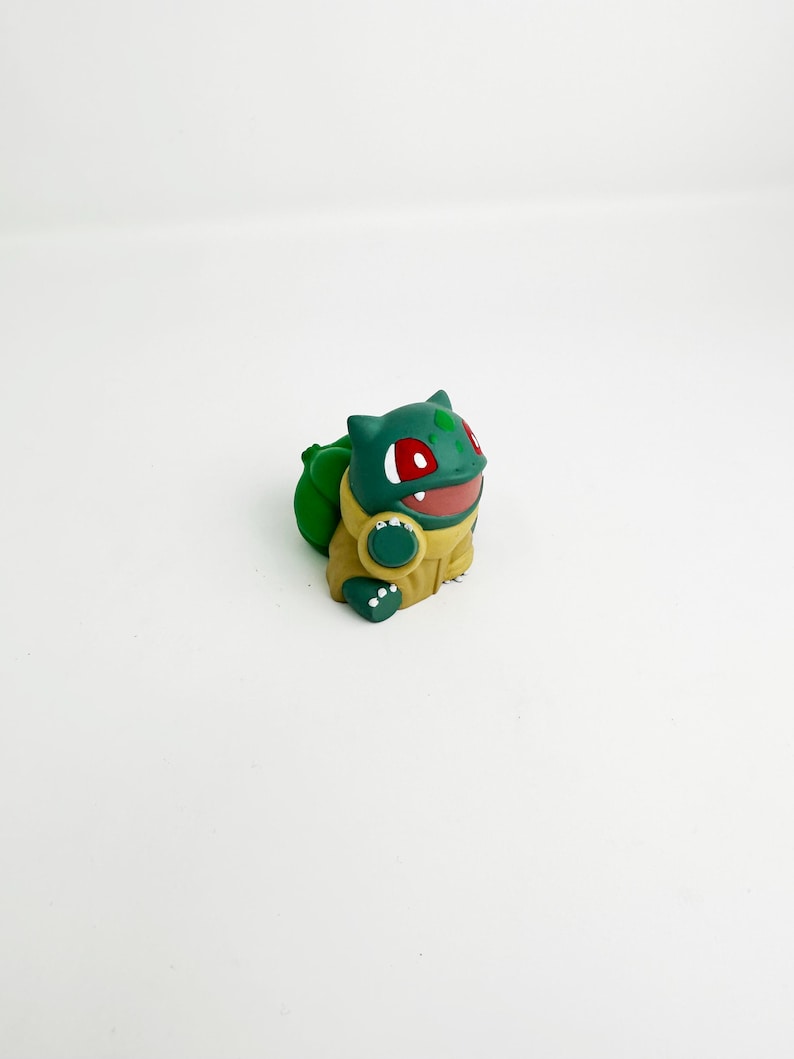 May include: A green and yellow Bulbasaur figurine with red eyes and a smiling mouth. The figurine has a large green bulb on its back and is set against a white background. The Bulbasaur is a popular character from the Pokemon franchise.