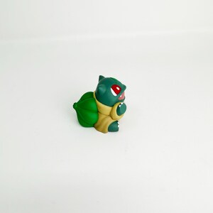 May include: A green Bulbasaur figurine with red eyes and a light brown body. The figurine has a large green bulb on its back and is set against a white background. The Bulbasaur is a popular character from the Pokemon franchise.