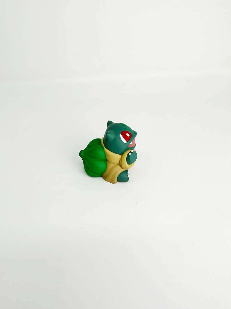 May include: A green Bulbasaur figurine with red eyes and a light brown body. The figurine has a large green bulb on its back and is set against a white background. The Bulbasaur is a popular character from the Pokemon franchise.