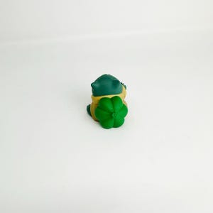 May include: A small, green and yellow toy frog with a green flower on its back. The frog has a dark green head and a yellow body. The flower is a bright green color. The toy is sitting on a white surface.