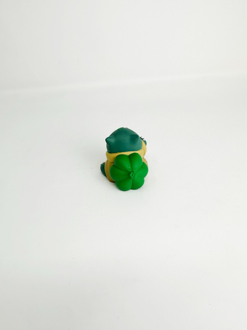 May include: A small, green and yellow toy frog with a green flower on its back. The frog has a dark green head and a yellow body. The flower is a bright green color. The toy is sitting on a white surface.