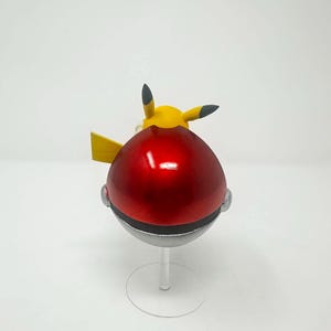 以下が含まれることがあります： 赤いと銀色のポケモンボールで、黄色いピカチュウのフィギュアが上にあります。ボールは光沢があり丸く、銀色のバンドと透明なスタンドが付いています。ピカチュウは黒い先端の耳と黄色の尾を持っています。